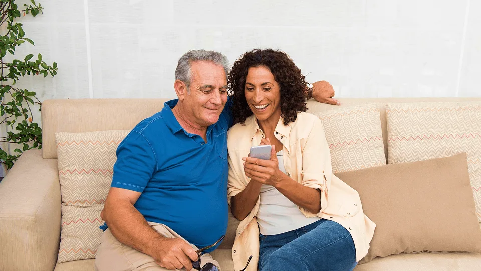 Casal sentado no sofá sorrindo enquanto confere informações no celular.