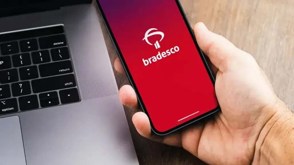 Mão segurando um celular com o logo do Bradesco ao lado de um notebook.